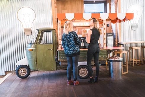 Image of two female colleagues getting a cup of coffee and talking next to a tuk tuk with a coffee machine on it. 
